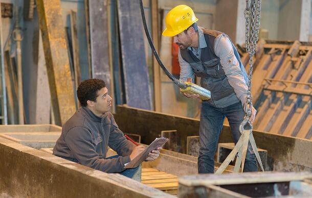 Two workers in a manufacturing facility communicate while inspecting equipment. One man, seated and holding a tablet, listens to the other, who is wearing a yellow hard hat and holding a control device attached to a suspended load.