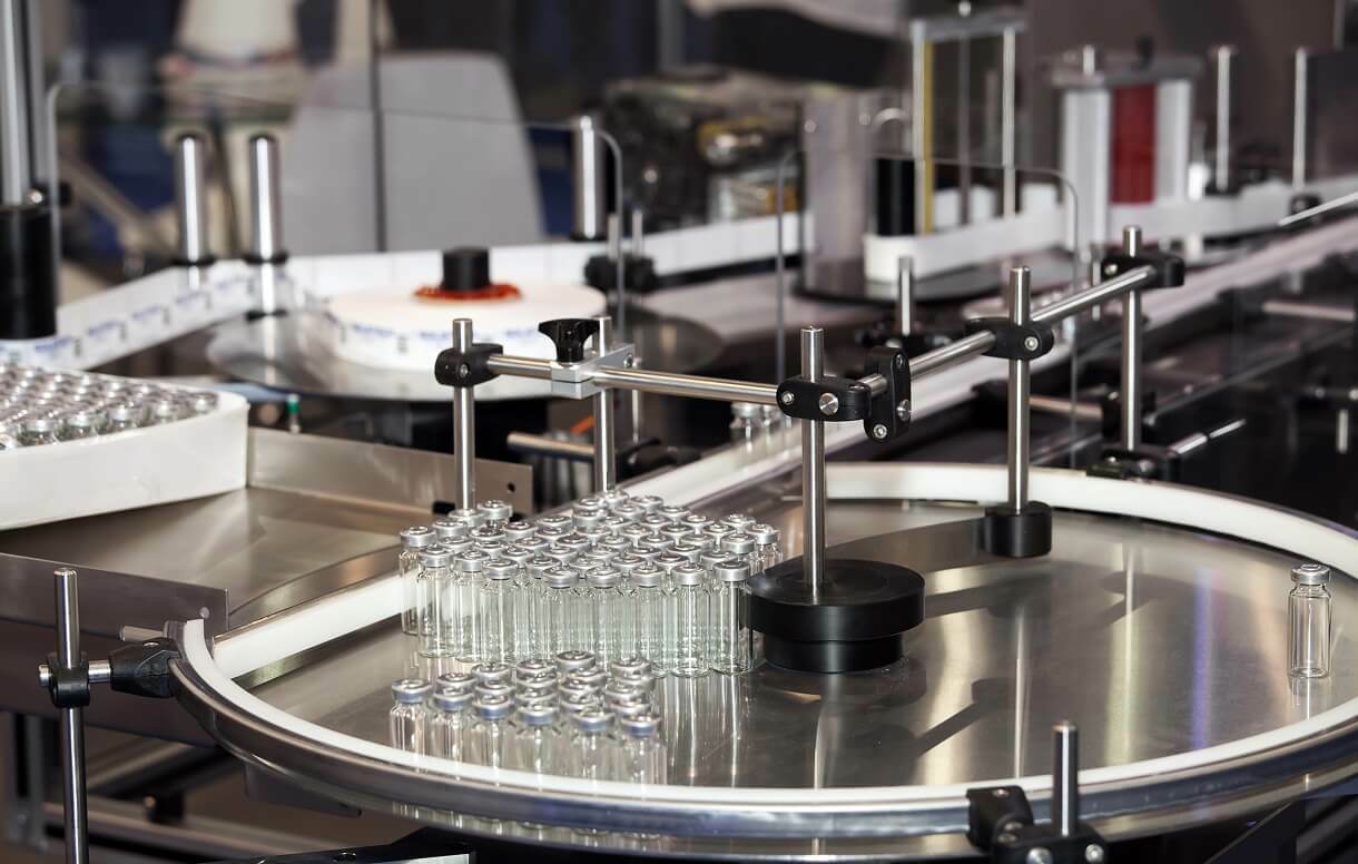 Automated pharmaceutical production line with glass vials moving along a conveyor system for filling and packaging.